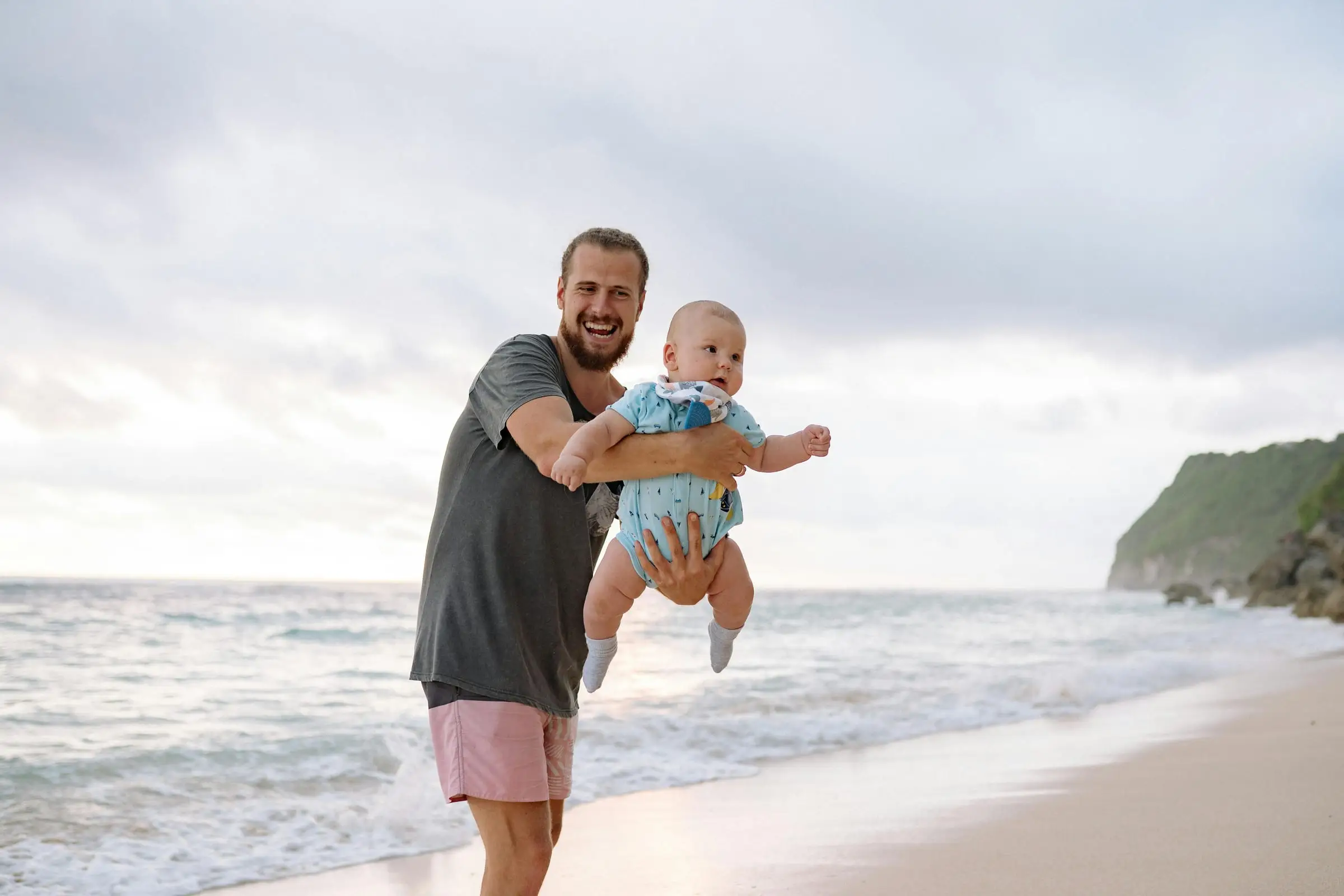 Ayah mengangkat bayi di pantai dengan tebing di belakang
